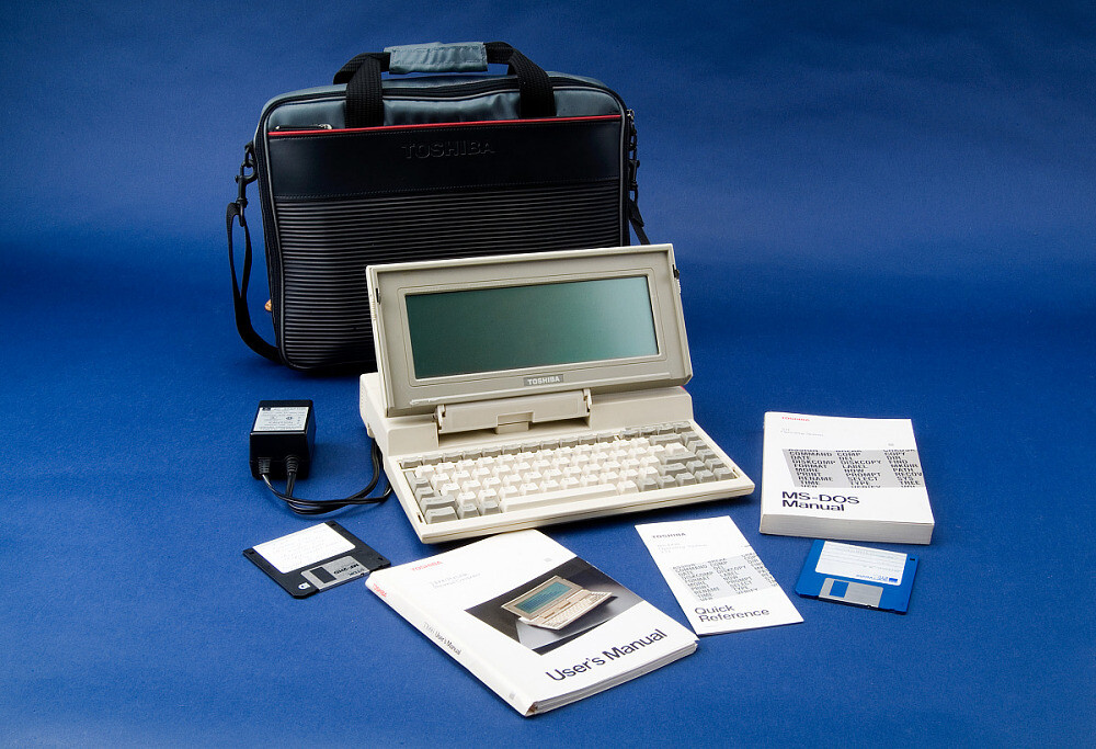 Foto de um portátil Toshiba T1000 com maleta, manual e disquetes, sobre um fundo azul. 
