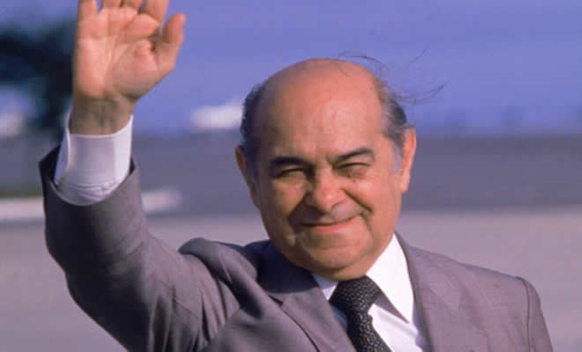 Foto de Tancredo Neves, de terno e gravata, em gesto de despedida