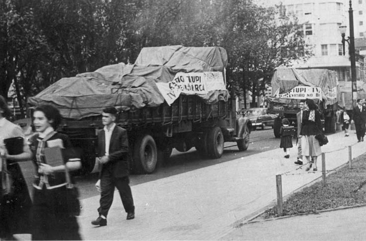 Foto em preto e branco de caminhões levando os equipamentos da TV Tupi, com faixas comemorativas.