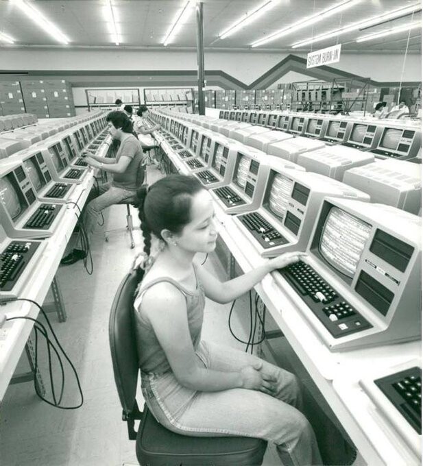 Foto em preto e branco de dezenas de computadores TRS-80 em bancadas, com uma operadora em primeiro plano, e mais algumas pessoas ao fundo. Uma placa presa ao teto diz: “SYSTEM BURN-IN“.