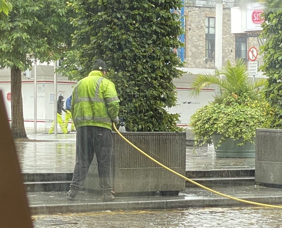 Foto de um homem na chuva, com um colete de alta visibilidade, molhando uma planta com uma mangueira