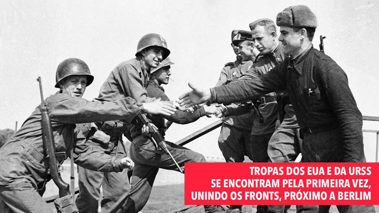 Foto em preto e branco de militares apertando as mãos, com legenda: tropas dos EUA e da URSS se encontram pela primeira vez, unindo os fronts, próximo a Berlim