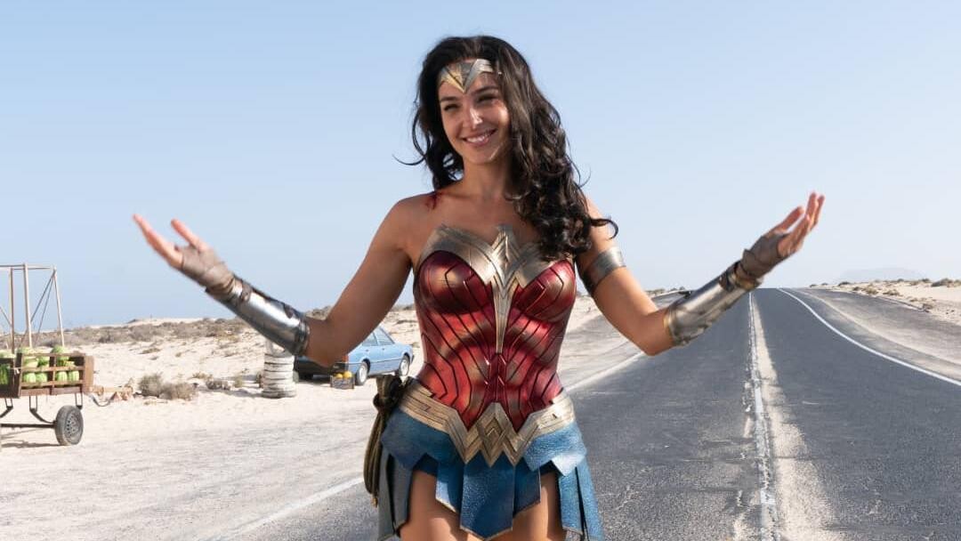 Gal Gadot, caracterizada como Mulher Maravilha, sorrindo de braços abertos no meio de uma estrada asfaltada