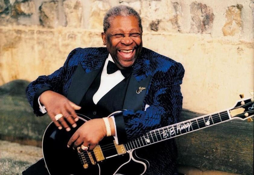 B. B. King, de traje de gala azul, com gravata borboleta, e com sua característica guitarra em mãos