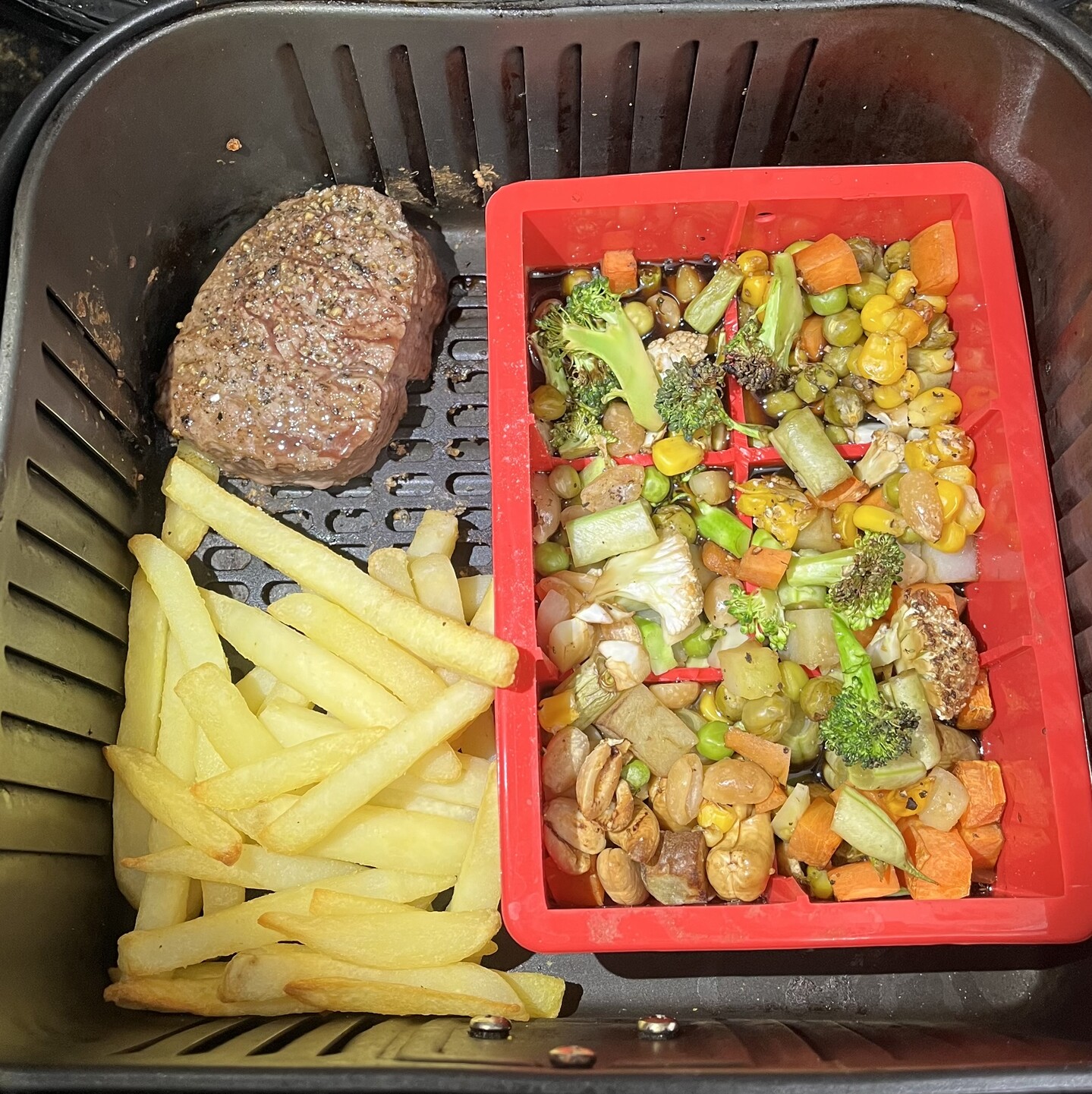 Cesto da airfryer contendo um bife com sal e pimenta do reino, batatas fritas e uma forma de silicone com os legumes picados e bastante shoyo