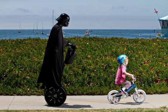 Darth Vader, em um patinete motorizado, passeia com uma criança em uma bicicletinha, num cenário praiano
