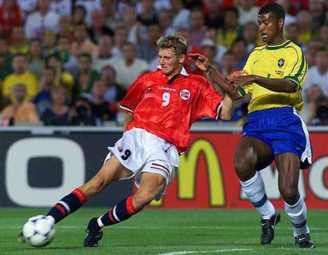 Centro-avante da Noruega, de uniforme vermelho, dribla jogador da seleção brasileira de futebol masculino.