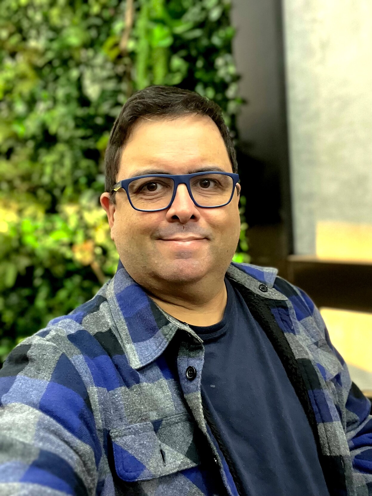 Homem adulto, de óculos e blusa de flanela, olhando para a câmera, com folhagem ao fundo