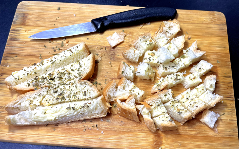 2 fatias de pão, com manteiga e ervas já aplicados, em processo de corte na tábua