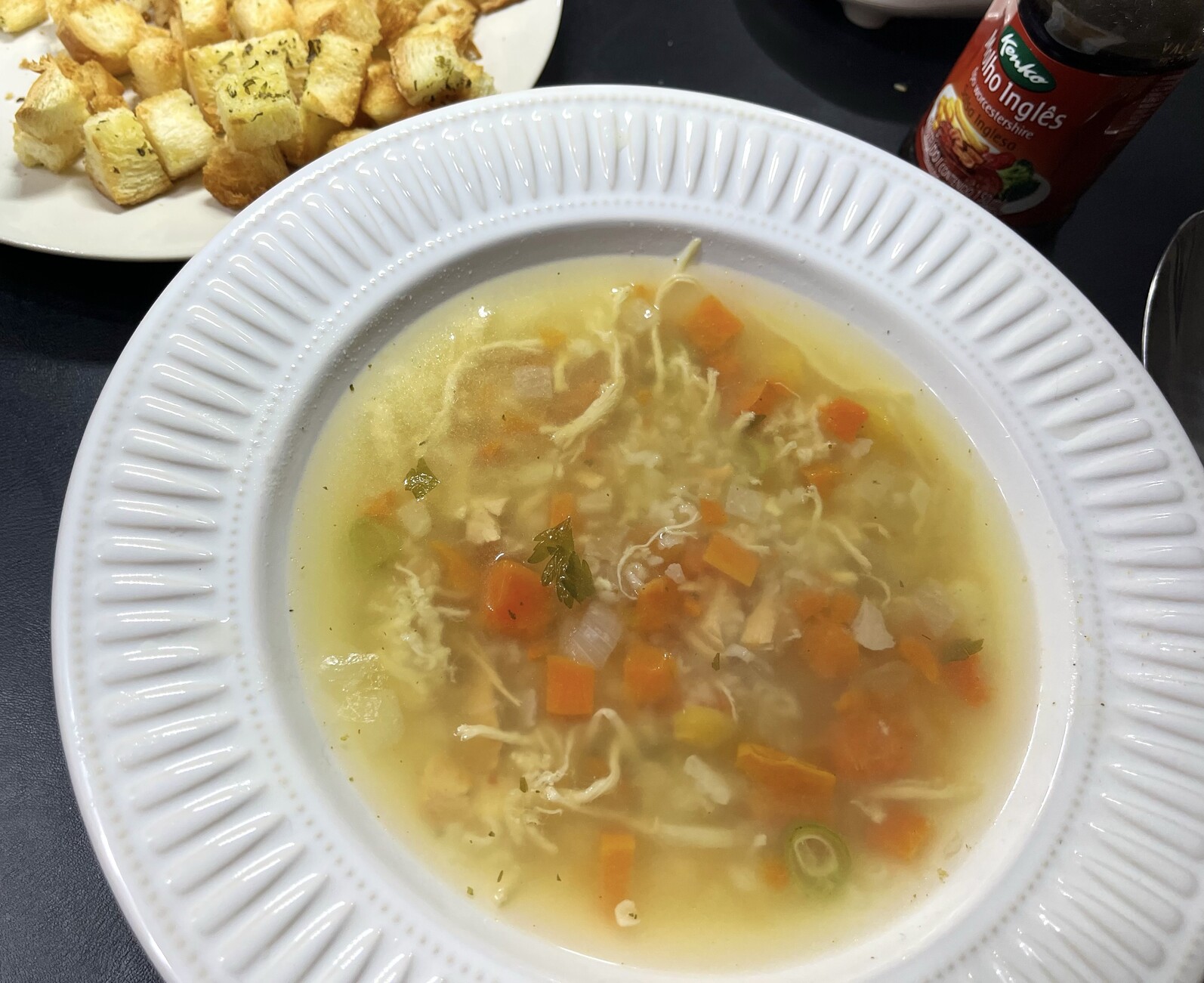Um prato de canja de galinha com legumes, ao lado de uma travessinha com torrada em cubos e de um frasco de molho Worcestershire 