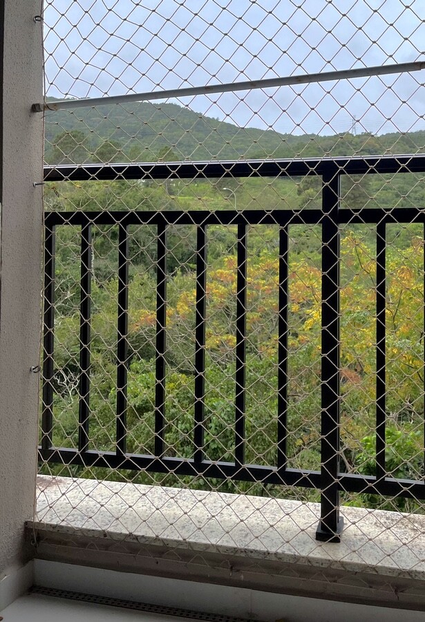 Vista olhando da sacada, para o mato e o morro, com uma tela de proteção e um guarda-corpo no caminho