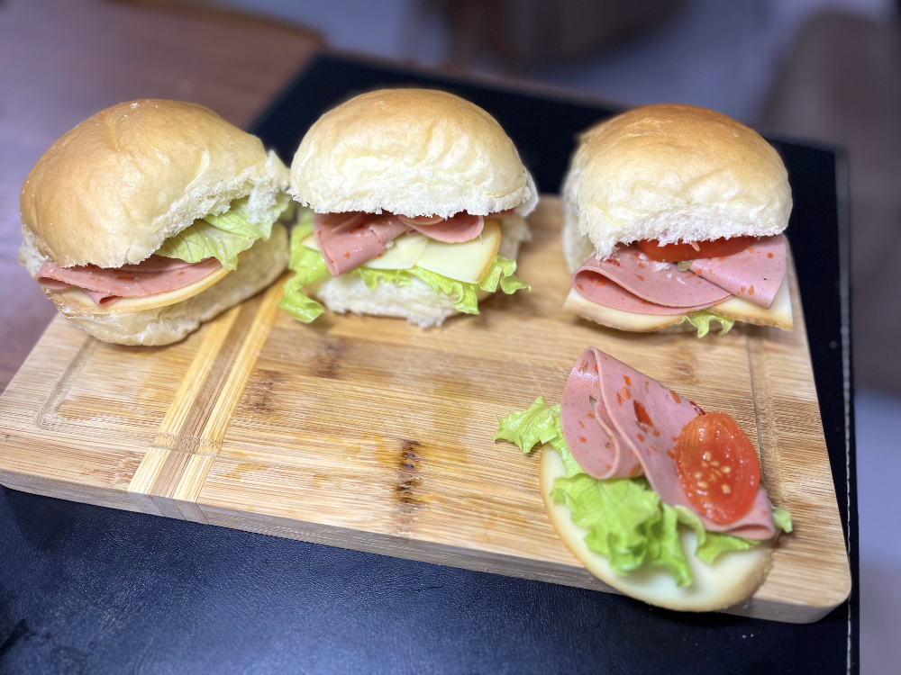 3 sanduíches de pãezinhos caseiros artesanais, com salsichão fatiado, provolone fatiado e alface e tomate cenográficos, em uma tábua decorada com o recheio de um deles