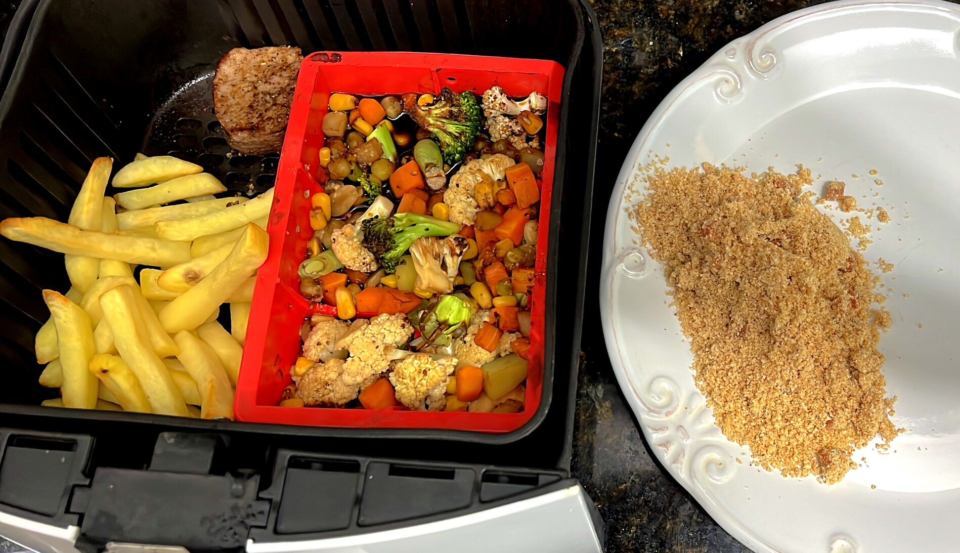 Airfryer aberta contendo batatas fritas, um bifinho bem temperado, e uma forma de silicone contendo legumes com bastante shoyo. Ao lado, o prato com a farofa já servida. 