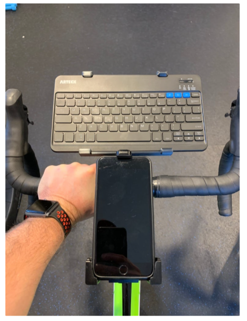 Mão segurando guidão de uma bicicleta de corrida, no qual estão instalados um celular e um teclado
