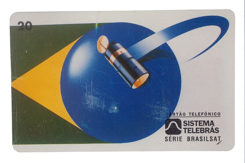 Cartão telefônico da Telebrás com 20 créditos da série Brasilsat. Parte frontal com fundo na cor branca contendo formas geométricas nas cores verde, amarela e azul remetendo à bandeira do Brasil. Por cima, contém desenho de satélite Brasilsat B1, da segunda geração do Brasilsat. Por cima da imagem, inscrições e logomarca do Sistema Telebrás na cor preta. Parte posterior na cor cinza contendo informações ao usuário sobre o cartão, inscritas na cor preta.<br>