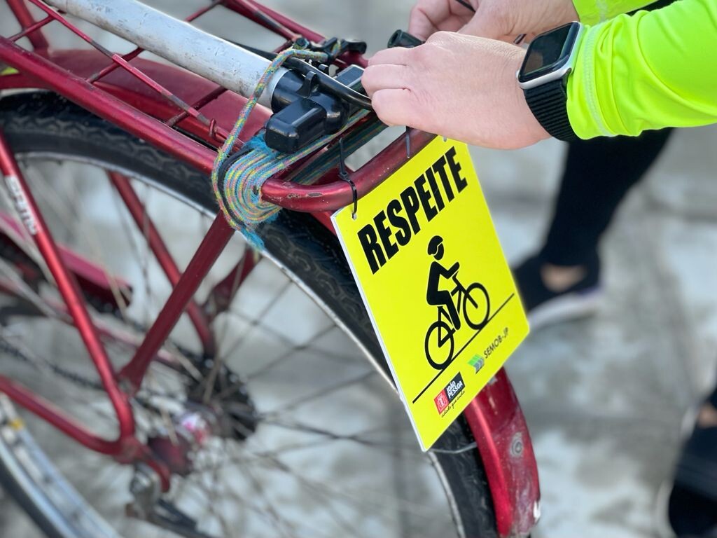 Ciclista afixa placa reflexiva, com a palavra RESPEITE, à traseira de sua bicicleta.