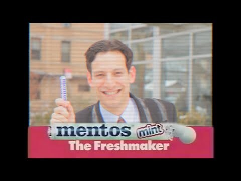 Jovem sorri segurando uma embalagem de Mentos em comercial de 1990