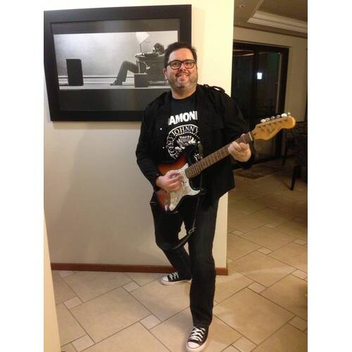 foto minha, de corpo inteiro, com jaqueta preta, calça jeans preta, tenis all star preto, segurando uma guitarra estilo stratocaster desplugada e sem usar a correia, e fazendo nela uma versão deslocada de um acorde típico da MPB, com uma reprodução de poster de Steve Steigman (1979) ao fundo.