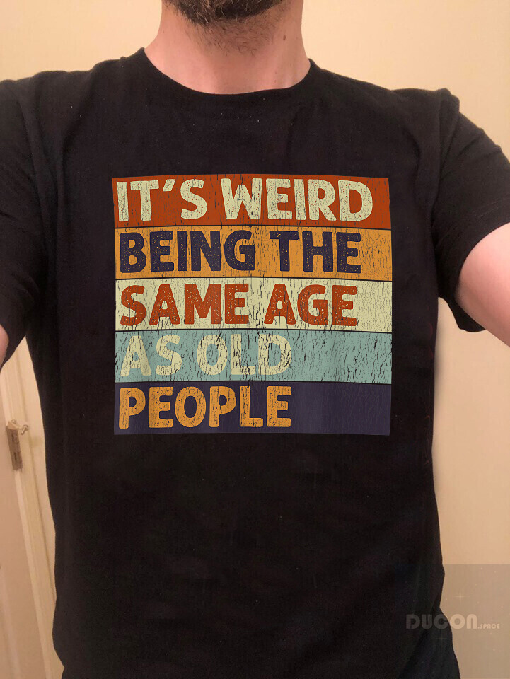 Torso masculino vestindo camiseta preta com estampa faux vintage com texto em cores de fundo variadas, dizendo: <br><br>IT'S WEIRD<br>BEING THE<br>SAME AGE<br>AS OLD<br>PEOPLE