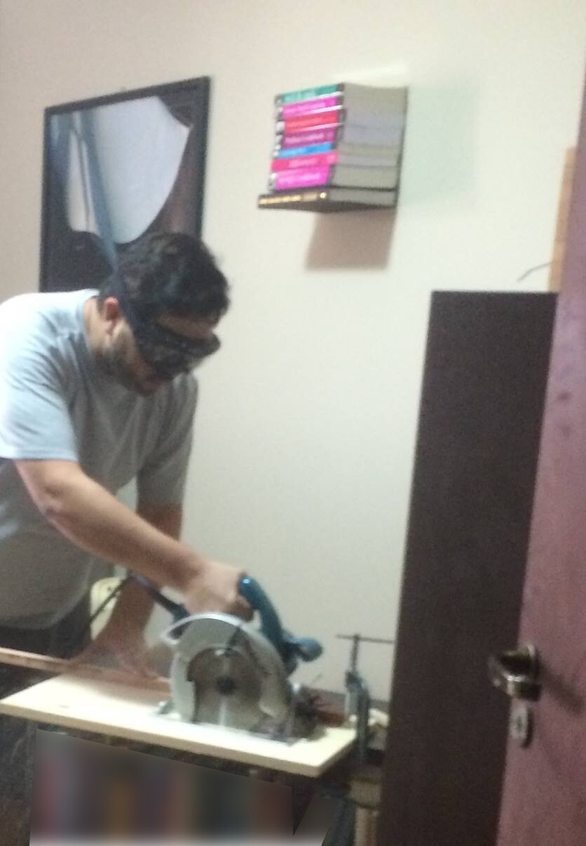 Foto minha segurando uma serra circular portátil, com óculos de proteção. Na parede atrás de mim, 8 livros montados como se estivessem flutuando encostados a ela.