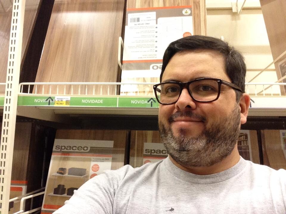 Selfie minha, com barba involuntariamente bicolor, camiseta cinza e óculos, em frente à prateleira de pranchas de compensado da Leroy Merlin 
