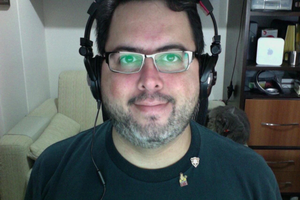 Selfie minha, de barba e óculos, com fones nos ouvidos e dois pins (do Linux Nacional e do Ubuntu) na camiseta azul marinho. Ao fundo, sofá e o gato Zi.