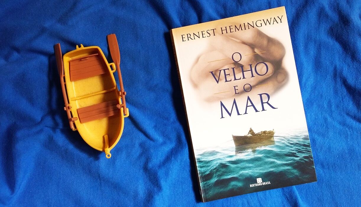 Exemplar de O Velho e O mar, sobre lençol azul e ao lado de um bote de brinquedo, amarelo, com 2 remos