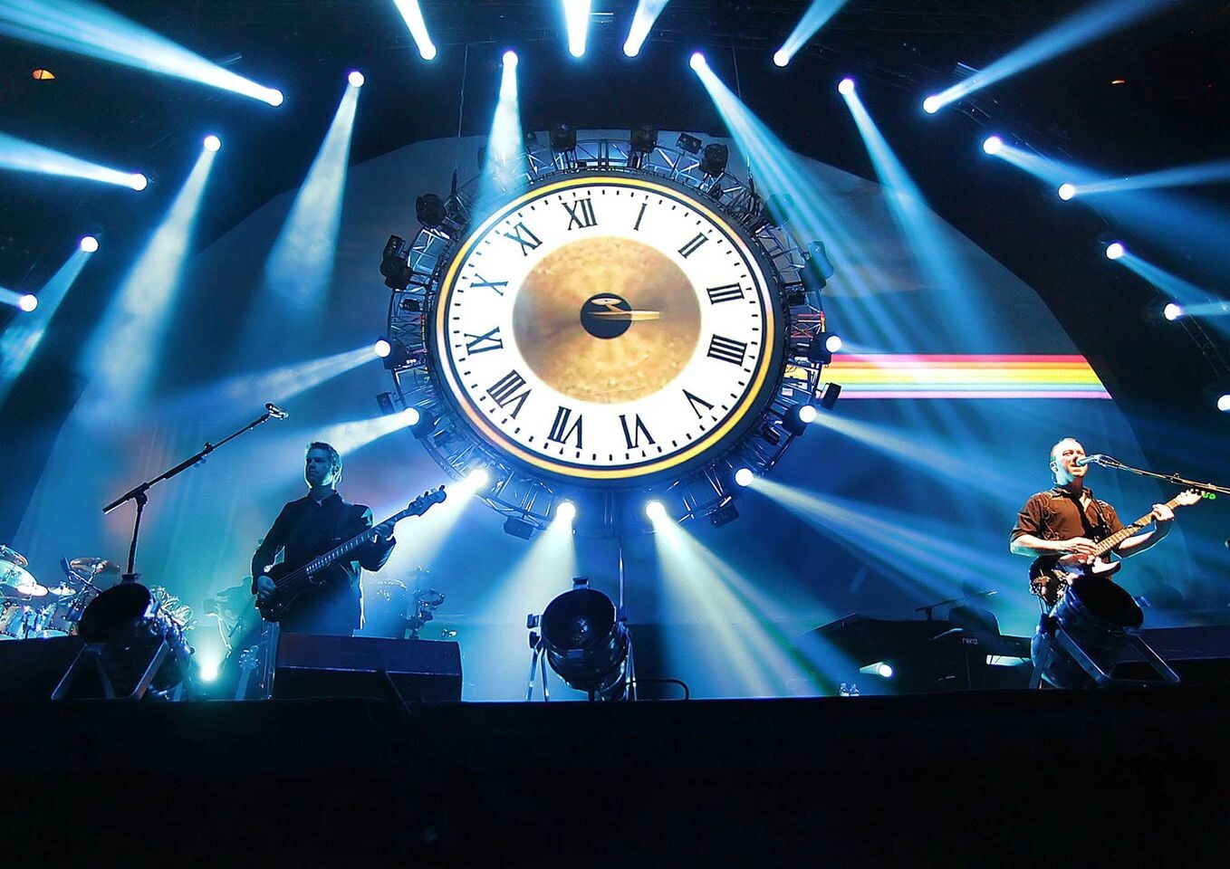 Baixista e guitarrista do Brit Floyd no palco, com um grande relógio antigo, sem ponteiros, decorando a cena