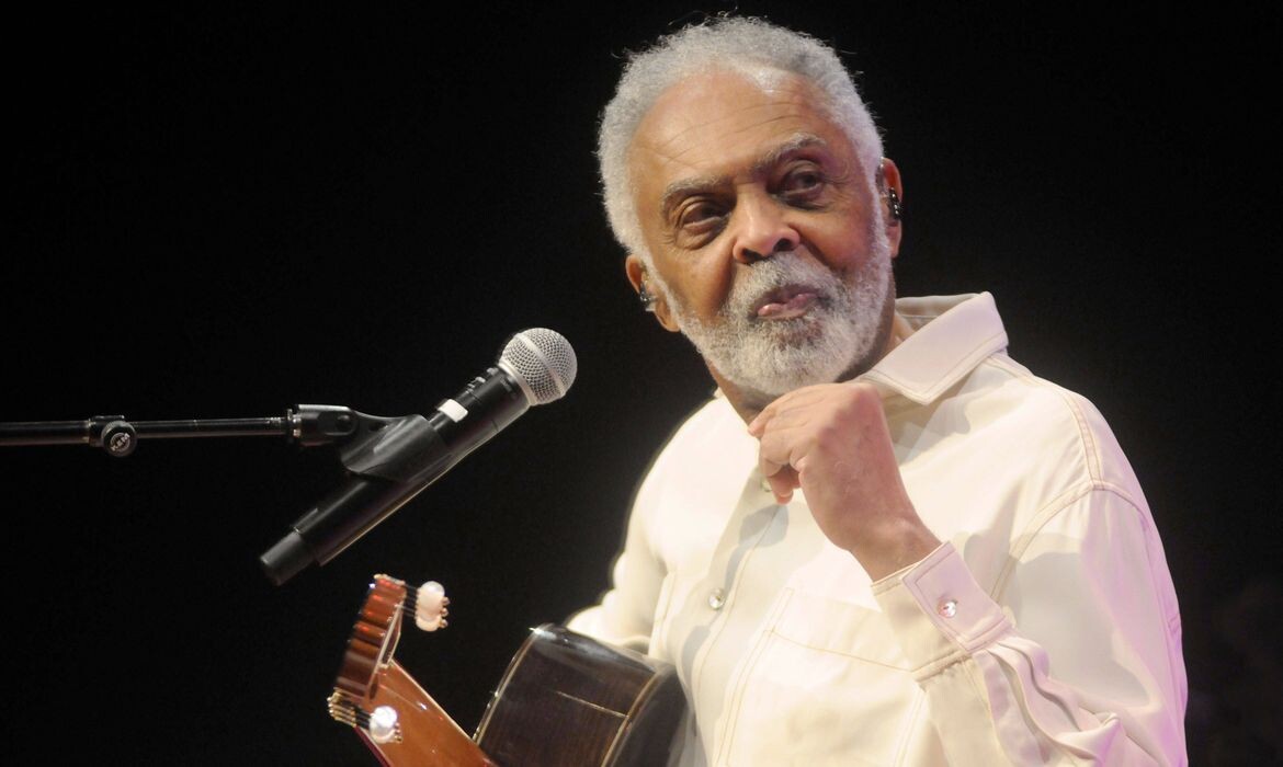 GIlberto Gil, de camisa branca, sorri no palco, com o violão em mãos