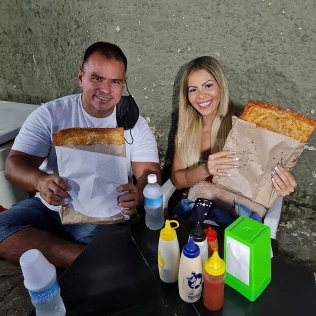 um casal, em mesa de lanchonete cheia de bisnagas de mostardas e maioneses, cada um segurando um pastel com dimensões de folha de papel A4, sorrindo para a câmera