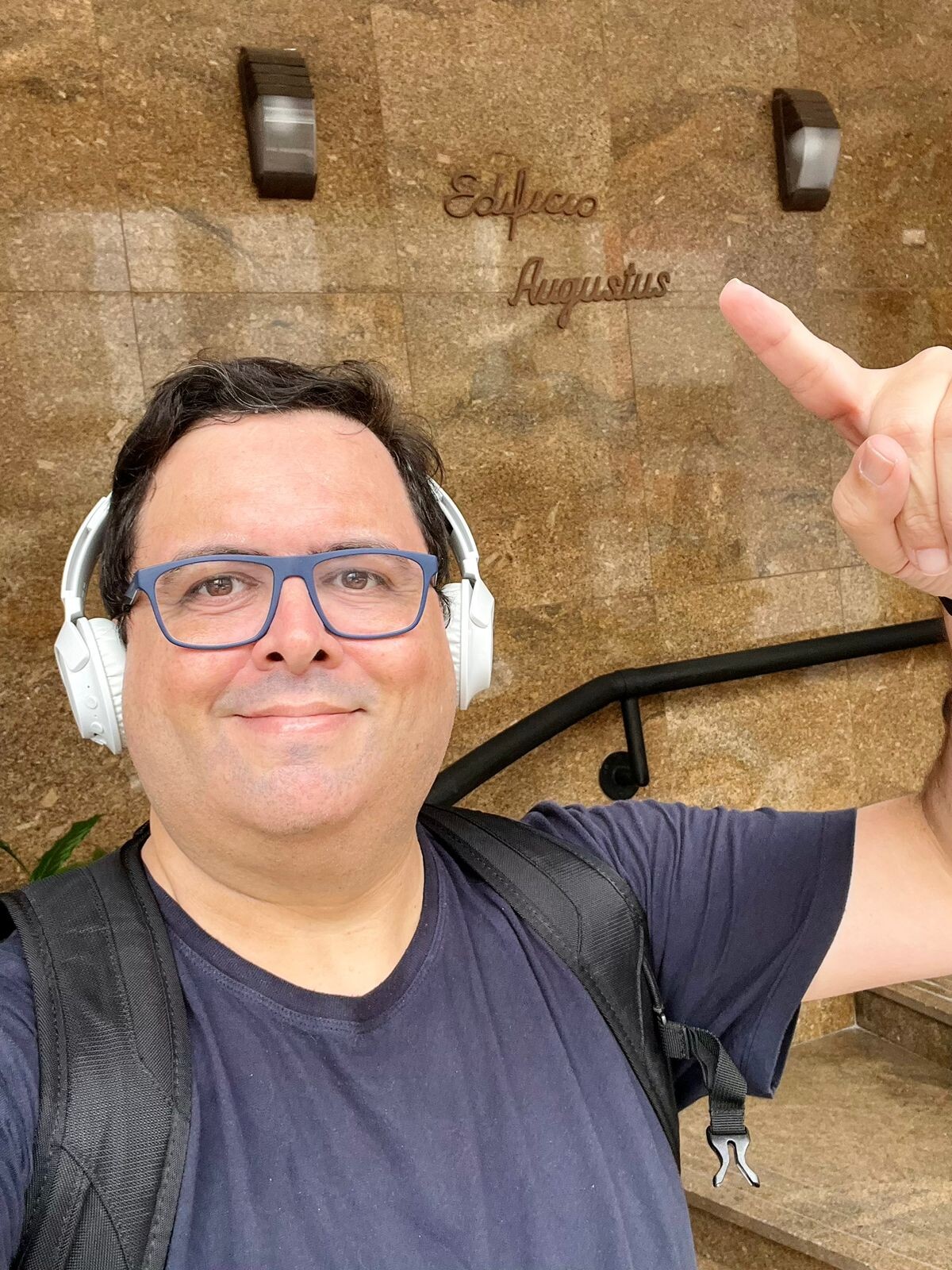Eu, olhando para a câmera, de camiseta azul, óculos ocm armação azul, fone de ouvido branco, e apontando para o nome do prédio: “Edifício Augustus“
