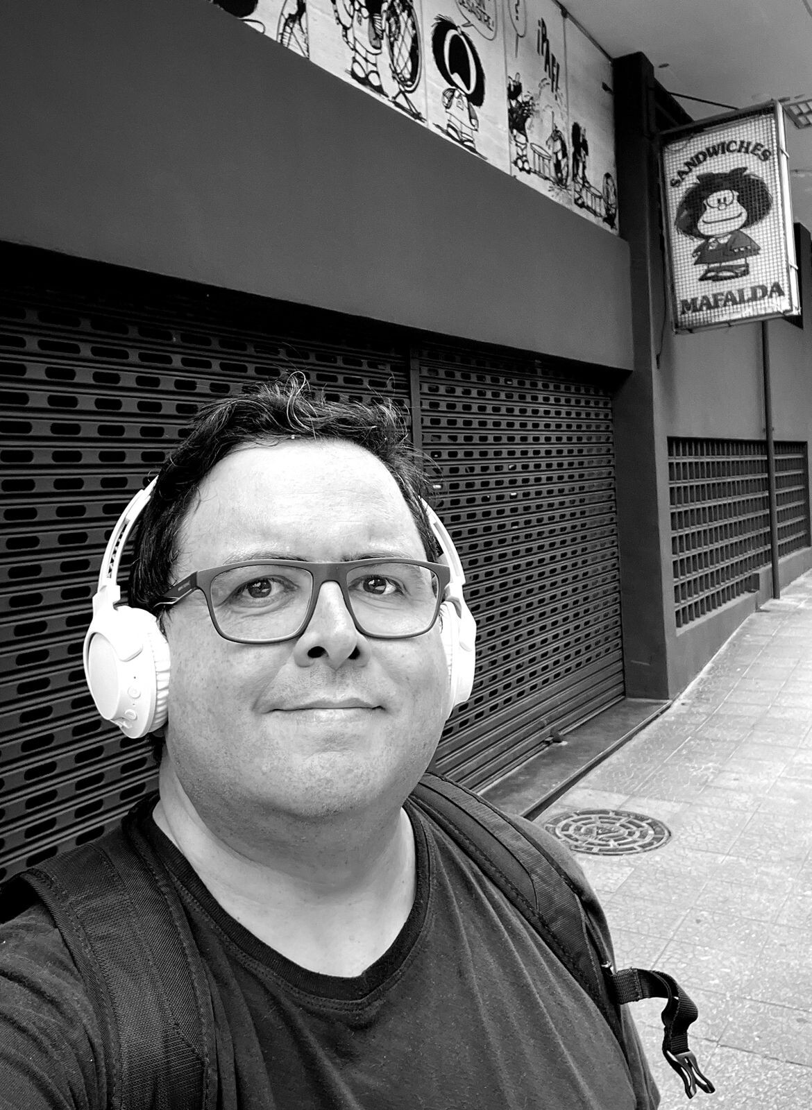 Foto em preto e branco, minha, olhando para a câmera, de óculos e fones de ouvido, em frente às portas fechadas da Sandwiches Mafalda