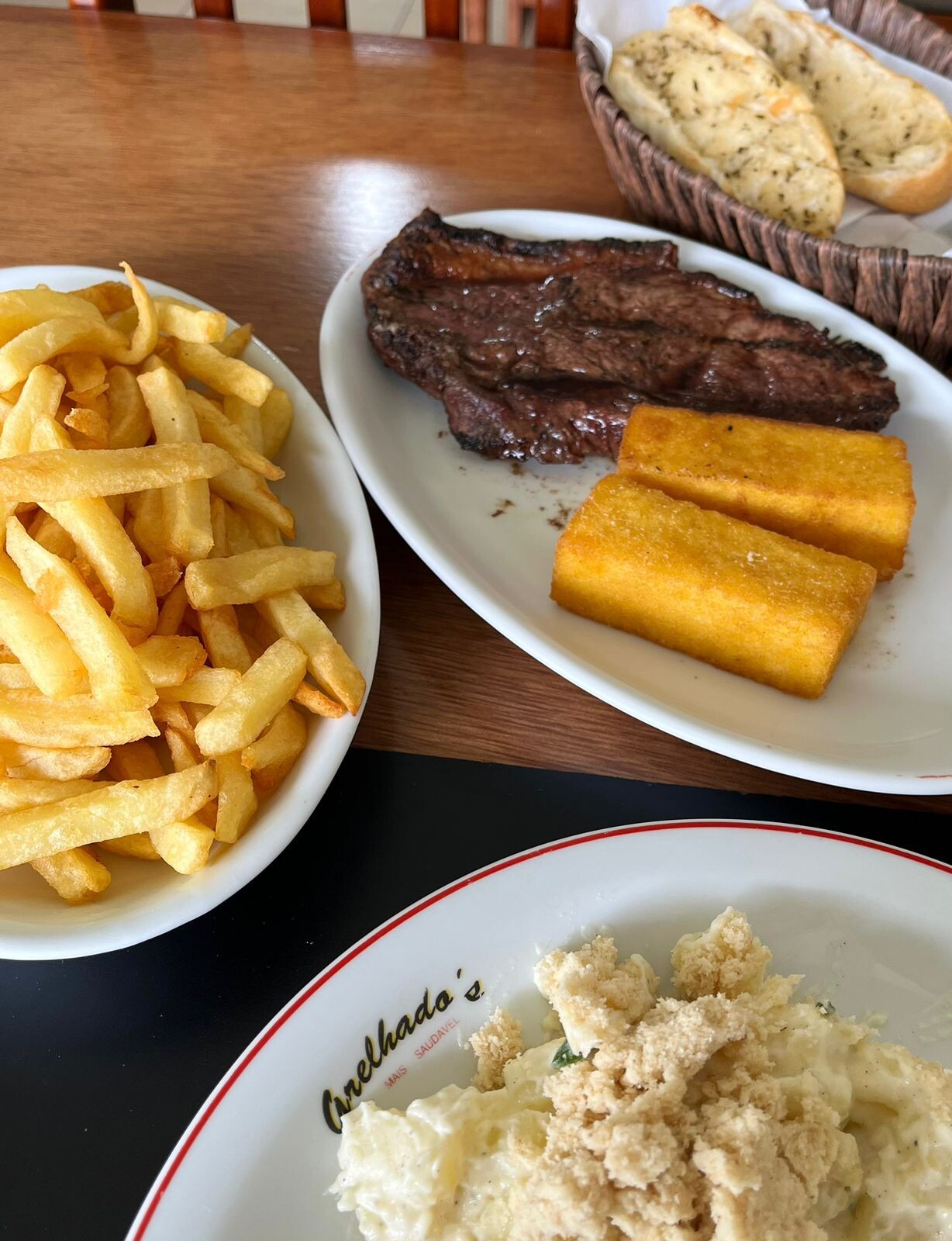 Duas polentas fritas, numa travessa com um bifão, ao lado de uma travessa de batatas fritas e de outra de pão com alho, posicionadas junto a um prato no qual há maionese e farofa, e se lê o nome “Grelhado's“