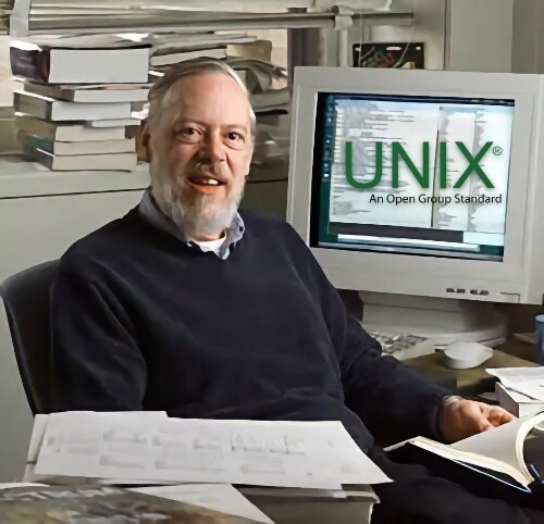 Dennis Ritchie, já na maturidade, folheia um manual, de costa para a tela de uma estação de trabalho onde se vê o logotipo Unix - an Open Group standard