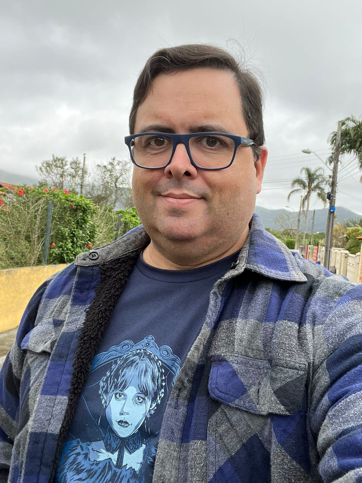 Foto minha olhando para a câmera, de blusa grunge desabotoada, camiseta azul da Wandinha (também em tons de azul) e óculos de armação azul. Ao fundo, plantas de um jardim, e o céu cinzento entre duas chuvas