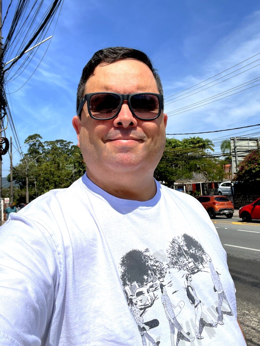Foto minha na margem da rodovia, com o céu azul como fundo, de óculos escuros com grau e camiseta branca com estampa reproduzindo, com personagens skatistas, a capa do disco Abbey Road, dos Beatles