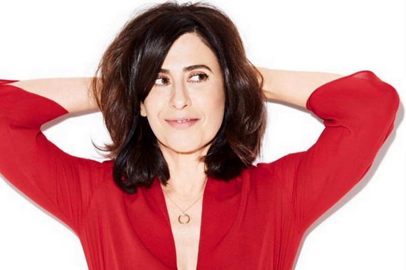 Fernanda Torres, trajada em vermelho e com as mãos na cabeça, sorri para a câmera e olha de soslaio