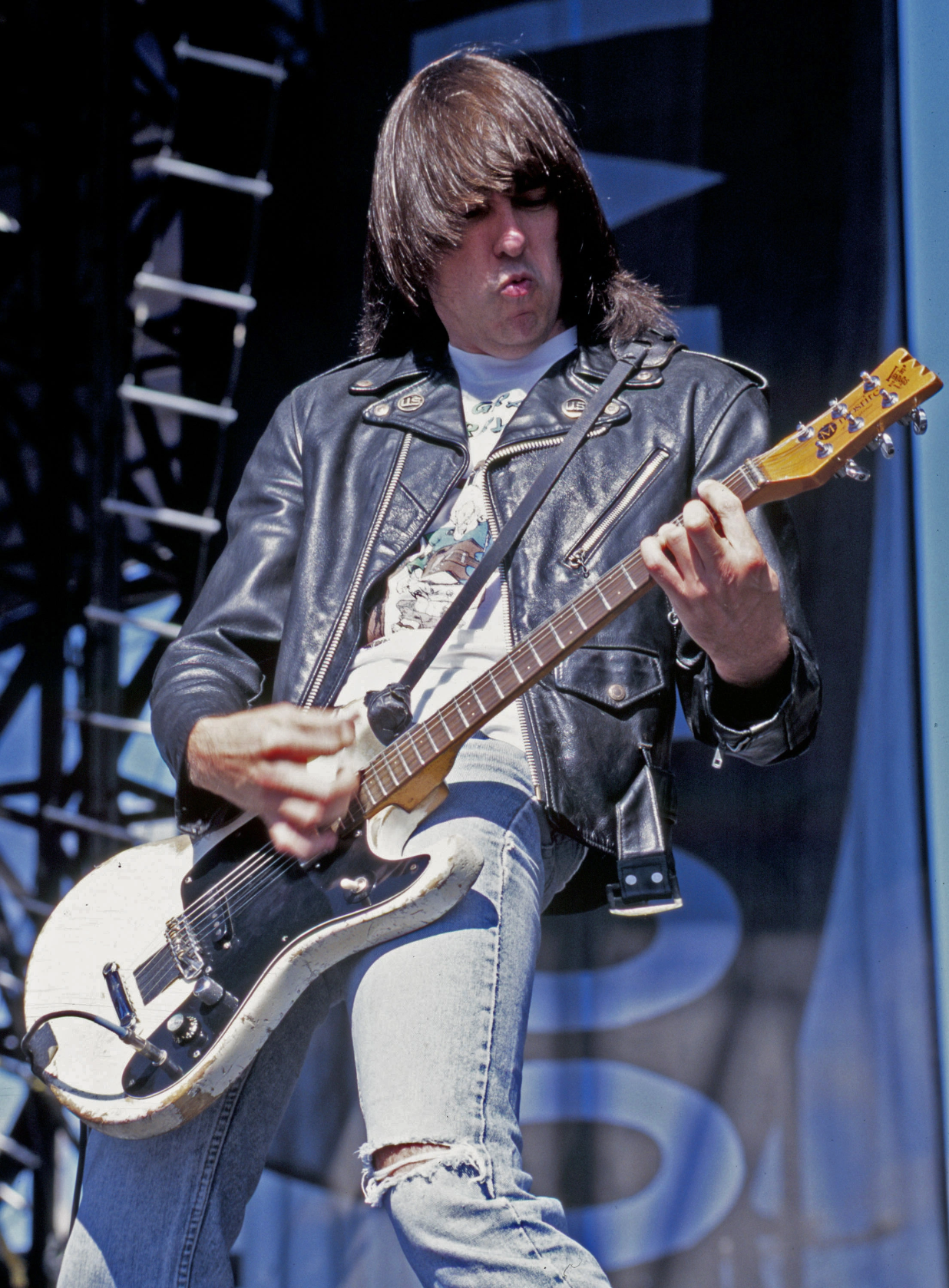 Johnny Ramone no palco, de jaqueta preta, com a guitarra