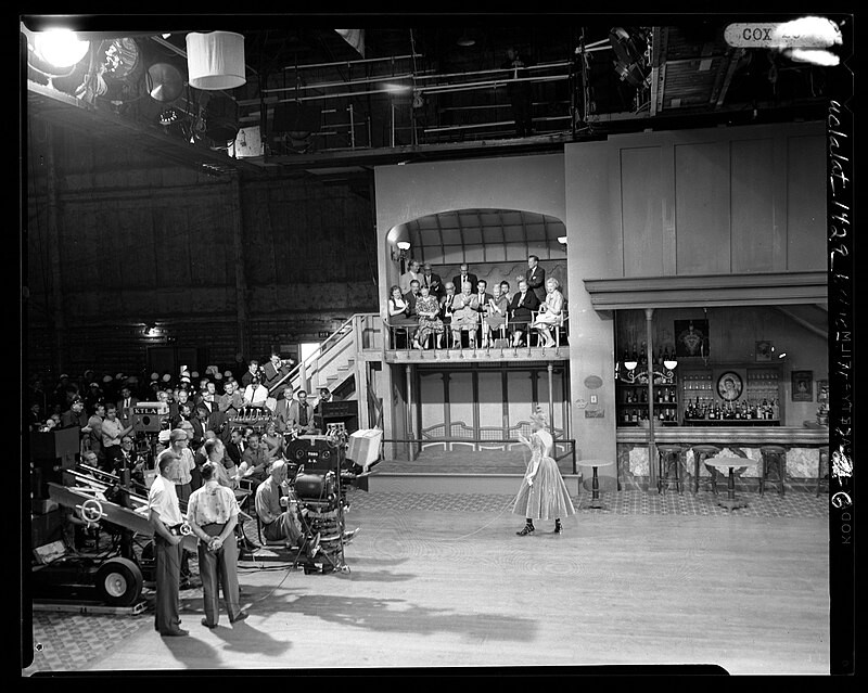 No dia seguinte, Khrushchev assiste às filmagens do filme “Can-Can“, que seria lançado em 1960. As pessoas no camarote , da esquerda para a direita, são: a filha do premier, Rada, o filho Sergei (atrás de Rada), a Sra. Eric Johnston, Spyras Skouras, o Primeiro-Ministro Khrushchev, a intérprete Sra. Buddy Adler (Anita Louise), a Sra. Khrushchev, a Sra. Skouras . Atrás de Alders está Nitkia, outra filha, Yulia, o embaixador Henry Cabot Lodge e Eric Johnston, presidente da Motion Picture Association.