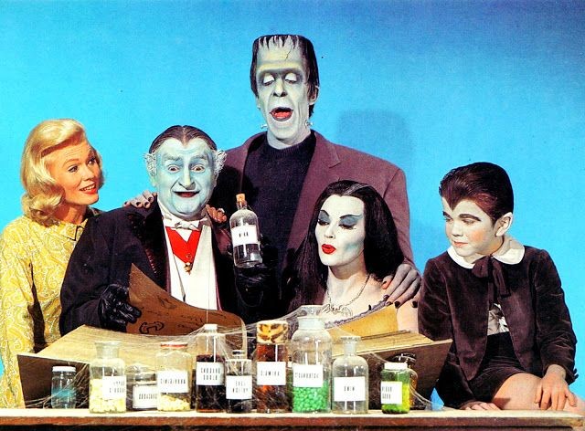 A família Munster reunida em foto colorida, folheando um grande livro, junto a uma prateleira de vidros de ingredientes