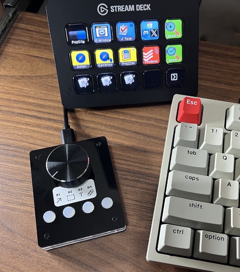 Foto da minha mesa mostrando o novo tecladinho, em cor preta, com um grande botão giratório e mais 4 botões circulares de cerca de 1cm de diâmetro cada. <br><br>Uma etiqueta indica a qual função do aplicativo de imagens cada botão foi associado. <br><br>Ao redor estão um Stream Deck indicando atalhos do navegador Safari, e a lateral de um teclado tradicional.