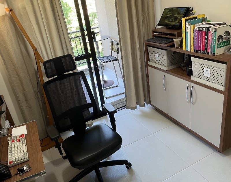 Foto do escritório mostrando uma estação de trabalho com teclado em bandeja removível e monitor pequeno, instalada no cantinho do tampo do móvel que até a véspera era a filial da biblioteca da casa, e no qual ainda há alguns livros de culinária e de tecnologia, em nome do histórico.