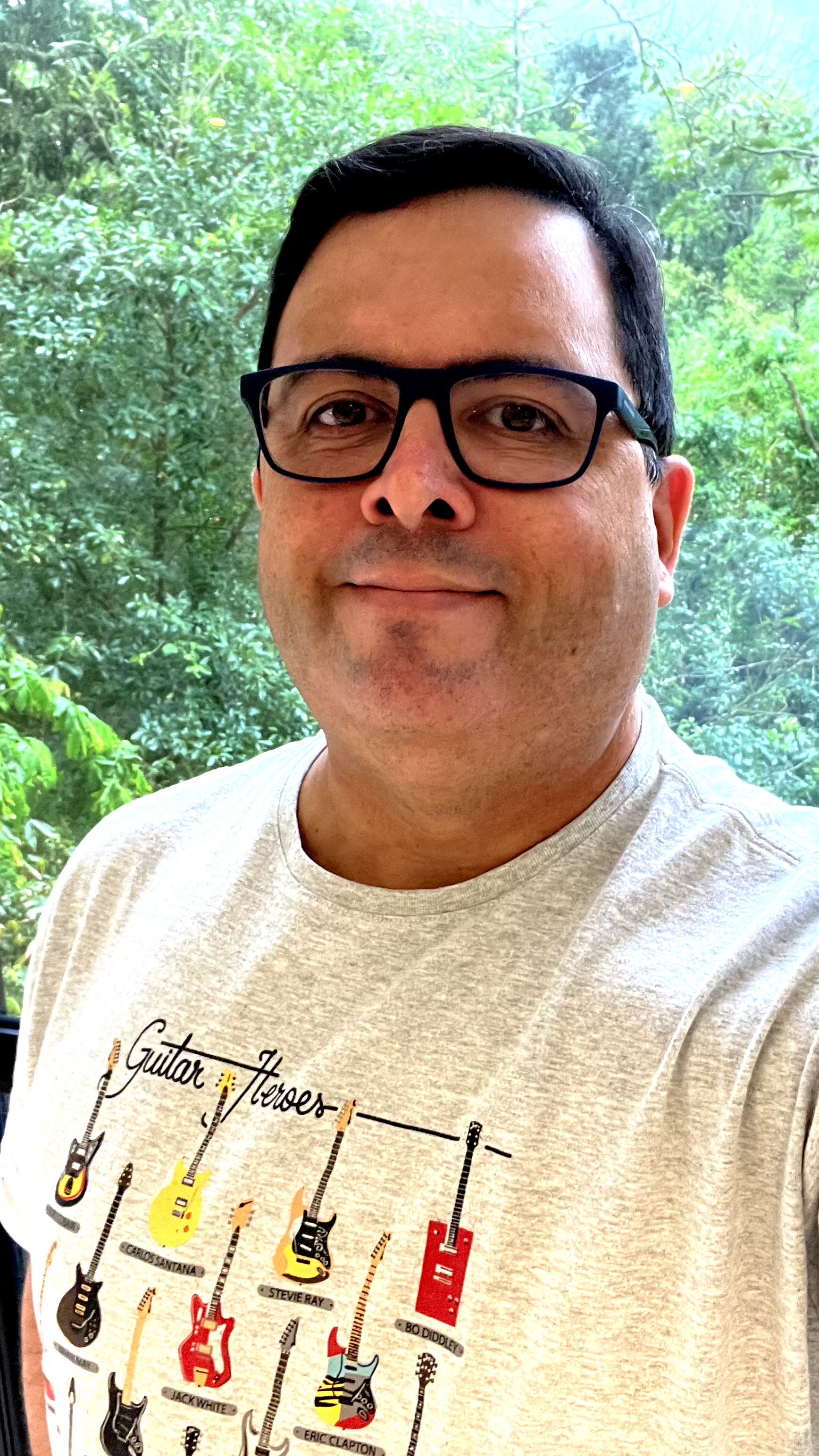 Foto minha de costas para a mata em meio à chuva, com oculos de armação azul e uma camiseta cinza com o titulo GUITAR HEROES e desenhos de guitarras de vários músicos, como Cobain, Brian May, Stevie Ray, Bo Diddley, Gilmour, Satriani e outros