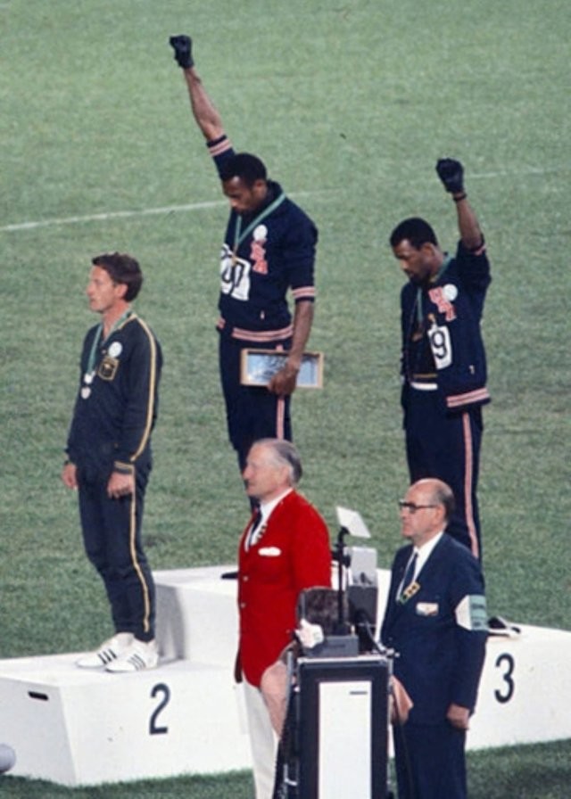 O medalhista de ouro Tommie Smith (centro) e o medalhista de bronze John Carlos (direita) mostrando o punho erguido, em saudação, no pódio após a corrida de 200m nos Jogos Olímpicos de Verão de 1968; ambos usam distintivos do Projeto Olímpico de Direitos Humanos. Peter Norman (medalhista de prata, à esquerda), da Austrália, também usa um distintivo do projeto, em solidariedade a Smith e Carlos.<br>