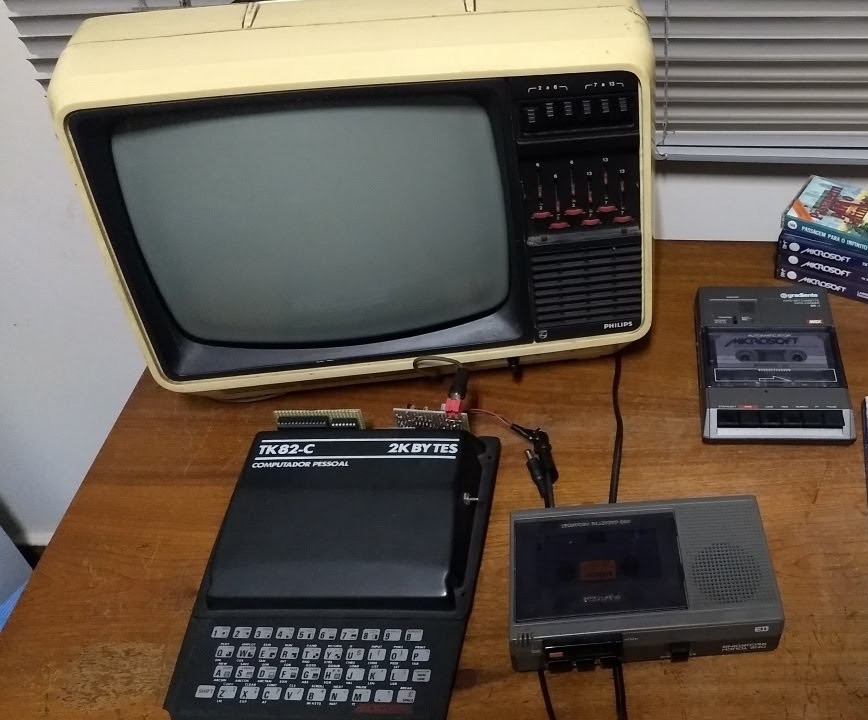Um TK82-C com expansões caseiras, sobre uma escrivaninha onde também estão uma TV em preto e branco, dois gravadores e algumas fitas K7
