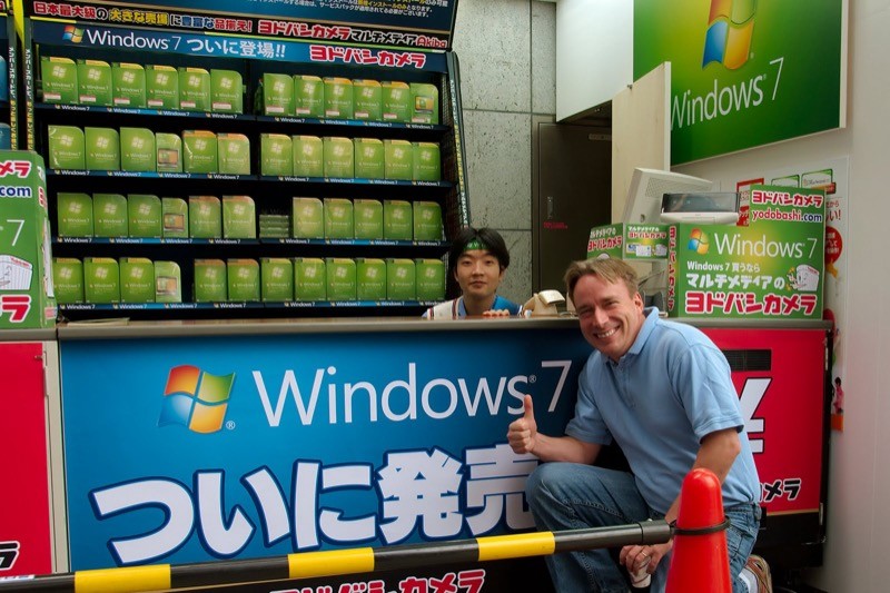 Linus Torvalds posa agachado e sorridente, fazendo sinal de positivo com o polegar direito, em frente a um stand do WIndows 7 no Japão
