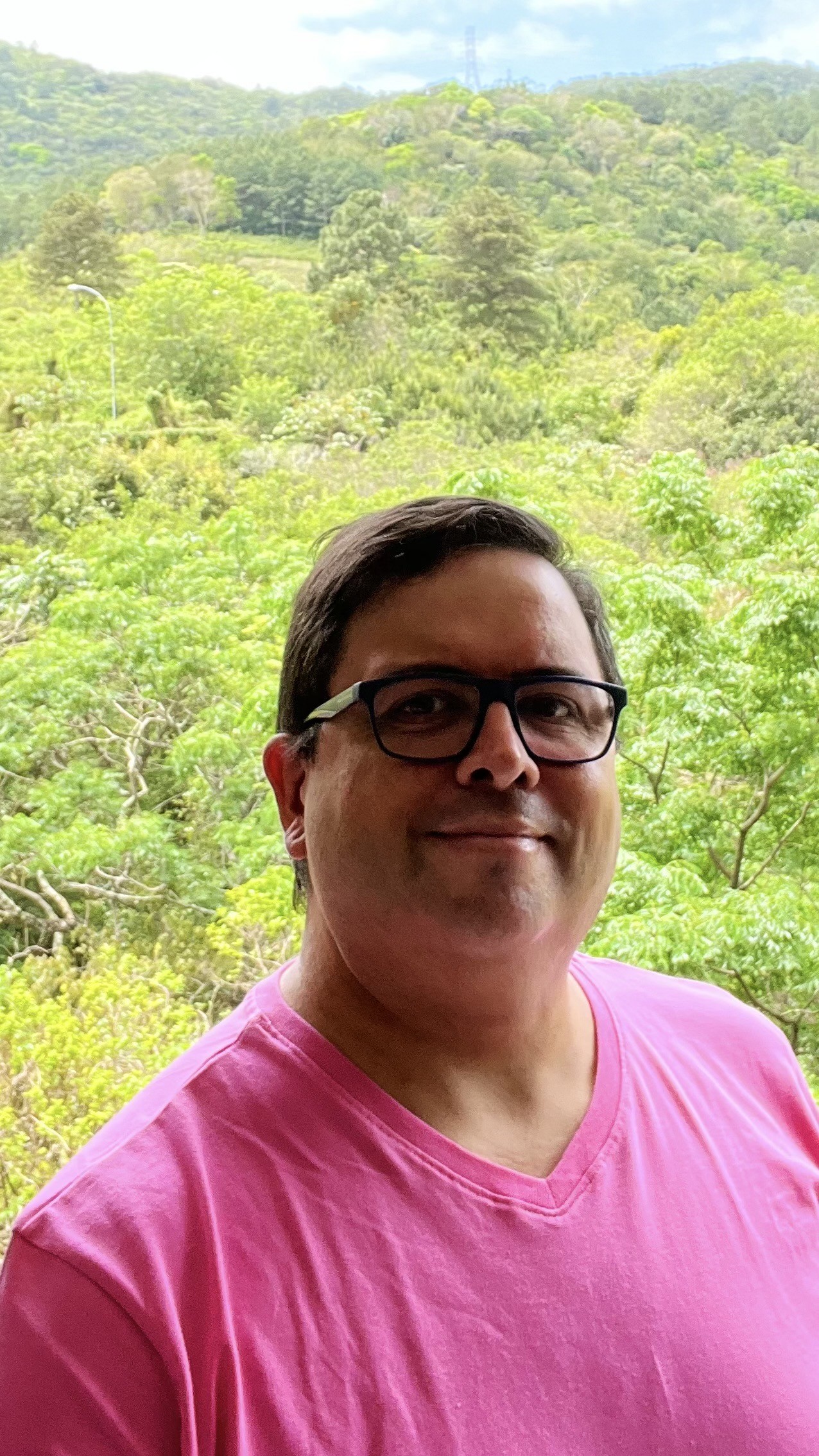Foto minha, de camiseta amarela e óculos de armação azul, sorrindo para a câmera, tendo a mata e o céu nublado ao fundo