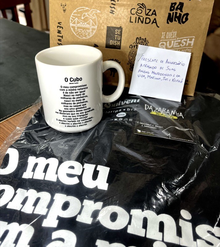 Camiseta e caneca com trechos da música O Cubo, do Dazaranha, e cartão onde se lê: <br><br>Presente de aniversário atrasado de suas amigas mastodônticas e da vida, Mariana, Tati e Keith!