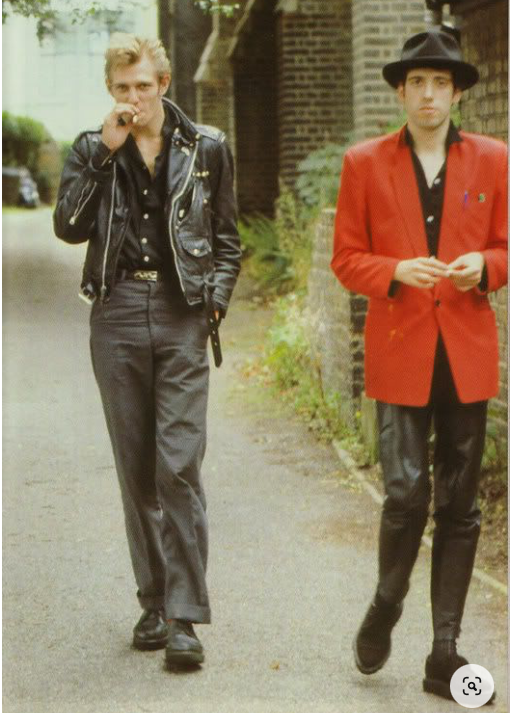 Dois integrantes do The Clash, de sapato, calça, terno e camisa social, sendo que um deles está de calça de couro, e o outro de jaqueta de couro.