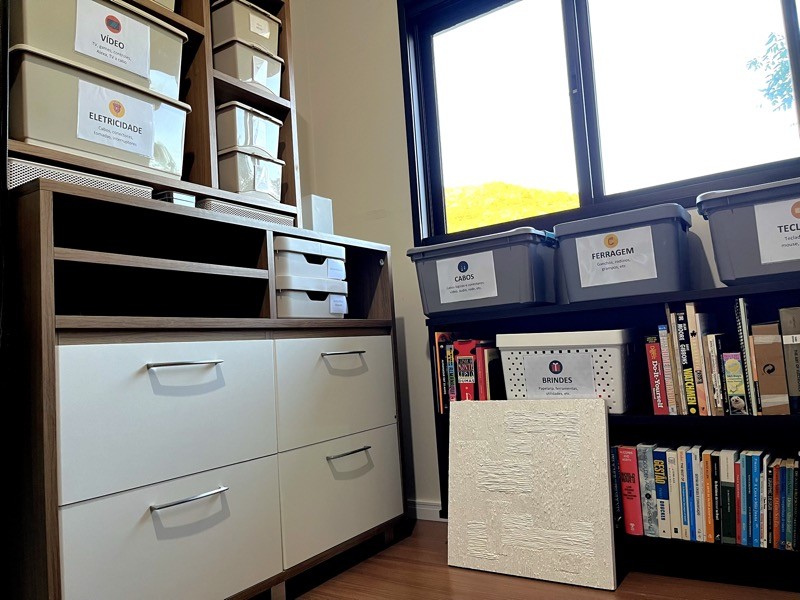 Uma cômoda e a estante de livros, abaixo de uma janela aberta, com um quadro da Feni Ehmke ainda esperando ser fixado na parede, e muitas e muitas caixas com etiquetas descrevendo seu conteúdo.